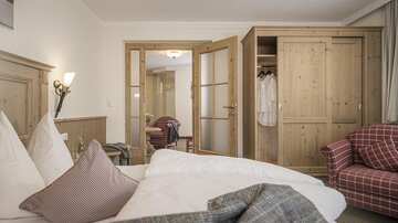 Ein gemütliches Zimmer mit einem Doppelbett und rustikalen Holzmöbeln. Im Hintergrund befindet sich ein Schrank und ein gemütlicher Sessel.