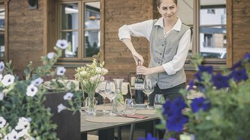 Eine junge Kellnerin deckt Tisch mit Gläsern und einer Flasche in einem Garten. Im Hintergrund sind Blumen und ein rustikales Holzgebäude zu sehen.