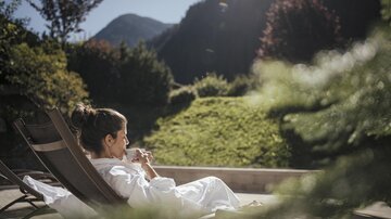 Eine Person in einem Bademantel entspannt auf einer Liege und genießt die Aussicht auf die grüne Landschaft. Im Hintergrund sind Berge und strahlender Sonnenschein zu sehen.
