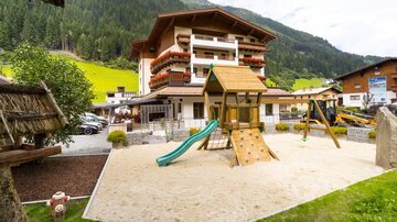 Ein Spielplatz mit Rutsche und Klettergerüst in einem ruhigen Bereich vor einem Gebäude. Im Hintergrund sind grüne Berge zu sehen.