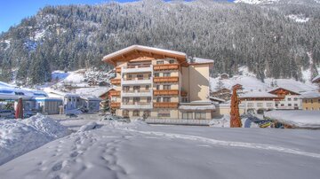 Ein schneebedecktes Dorf mit einem großen Gebäude im Vordergrund. Hinter dem Gebäude erheben sich schneebedeckte Berge unter einem klaren blauen Himmel.