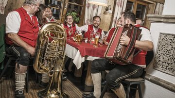 Eine Gruppe von Musikern in traditioneller Kleidung spielt in einem gemütlichen Gasthaus. Auf dem Tisch stehen Getränke und es herrscht eine fröhliche Atmosphäre.