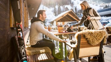 Ein Paar sitzt auf einer Terrasse in den Bergen, umgeben von schneebedeckten Hügeln. Die Sonne scheint, während die Frau dem Mann einen Skihelm reicht.