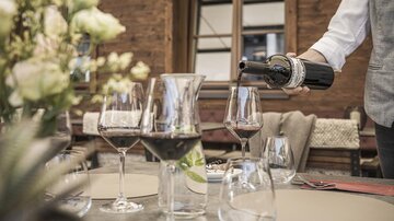 Ein eleganter Tisch mit Weingläsern und einer Karaffe. Jemand schenkt gerade Rotwein ein und es gibt eine Blumenarrangement im Hintergrund.