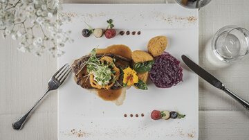 Ein kunstvoll angerichteter Teller mit Fleisch, buntem Gemüse und lila Rotkohl. Dazu ein Glas Wein und eine stilvolle Tischdekoration.