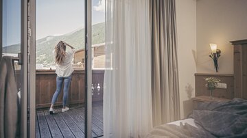 Eine Frau steht auf einem Balkon und schaut auf die Berge. Der Raum ist gemütlich und gut beleuchtet.