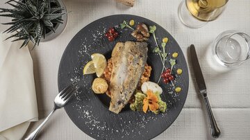 Ein schön angerichteter Teller mit gebratenem Fisch, Zitronenscheiben und buntem Gemüse. Dazu ein Glas Wein und eine Pflanze im Hintergrund.