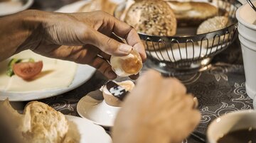 Ein Frühstückstisch mit verschiedenen Brötchen und einer Tasse Kaffee. Eine Hand bricht ein Brötchen auf, um es mit Aufstrich zu versehen.