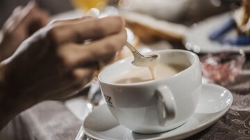 Eine Hand rührt in einer Tasse Kaffee auf einem Tisch. Im Hintergrund sind einige Snacks und Getränke zu sehen.