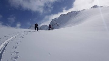 Zwei Personen gehen durch eine verschneite Landschaft. Der Himmel ist blau mit einigen Wolken.