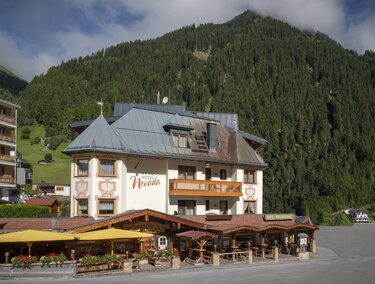 Ein charmantes Hotel mit einem großen Balkon und einer einladenden Terrasse. Im Hintergrund sind grüne Berge und eine bewölkte Himmel zu sehen.