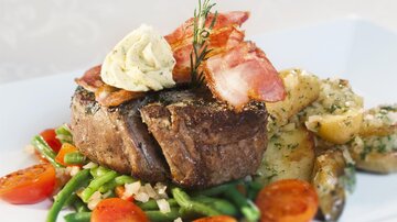 A delicious dish with a juicy steak, topped with bacon and herb butter. Served with green vegetables, halved tomatoes, and golden potatoes.