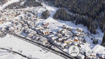 Eine malerische Winterlandschaft mit schneebedeckten Gebäuden und Bäumen. Im Vordergrund verläuft eine Straße durch das Dorf. | © seiblishof