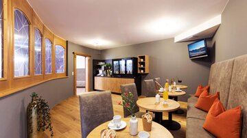 Ein gemütlicher Café-Bereich mit hellen Tischen und bequemen Sitzgelegenheiten. Auf den Tischen stehen Getränke und kleine Pflanzen.