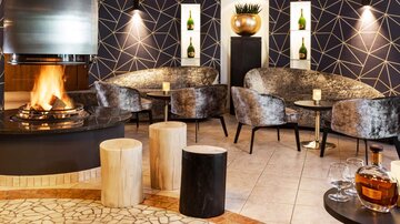 Ein eleganter Lounge-Bereich mit gemütlichen Sesseln und einem offenen Kamin. Die Wände sind modern gestaltet und die Atmosphäre ist einladend.