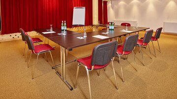 Ein moderner Konferenzraum mit einem großen Tisch und Stühlen. An der Wand hängen rote Vorhänge, und es gibt ausreichend Licht für Meetings.
