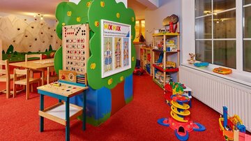 Ein bunter Spielraum mit einem grünen Spieltisch und verschiedenen Spielzeugen. Die Wände sind hell, und der Boden ist mit rotem Teppich ausgelegt.
