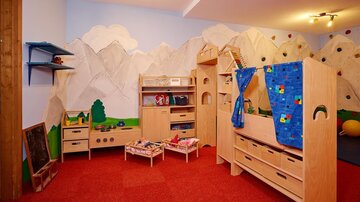 Ein kinderfreundlicher Spielraum mit Holzmöbeln und bunten Vorhängen. Die Wände sind mit Bergmotiven dekoriert, und der Boden ist mit rotem Teppich ausgelegt.