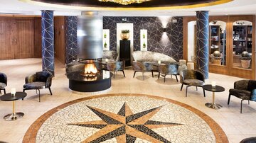 Ein eleganter Lobbybereich mit modernen Sesseln und einem zentralen Kamin. Der Boden ist mit einem auffälligen Mosaik in Form eines Kompasses gestaltet.