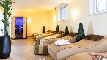 Ein entspannender Wellnessbereich mit bequemen Liegen und Pflanzen. Im Hintergrund ist eine Sauna zu sehen, die eine ruhige Atmosphäre schafft.