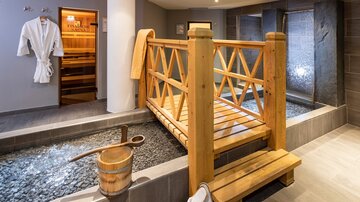Ein eleganter Wellnessbereich mit einem Holzsteg über einem Kiesbett. Im Hintergrund sind eine Sauna und entspannende Wasserfälle zu sehen.