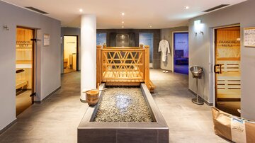 Ein moderner Wellnessbereich mit einem Wasserbecken in der Mitte. Umgeben von Saunen und entspannenden Elementen.