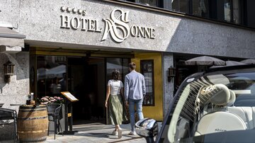 A modern hotel with a stylish entrance and elegant signage. A couple enters the hotel "Sonne," surrounded by an inviting atmosphere.