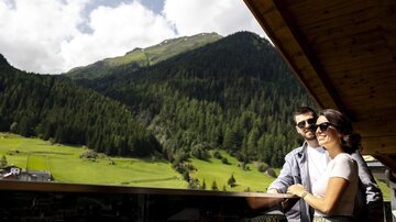A couple stands on a balcony and enjoys the view of the green mountains and forests. The sky is partly cloudy and the landscape is idyllic.