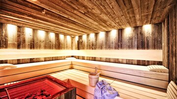 A modern sauna with bright wooden paneling and gentle lighting. In the center, there is a steaming container, and the benches invite you to relax.