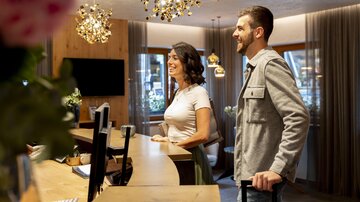 A friendly couple stands at the reception desk of a hotel. The environment is modern and inviting, with attractive lighting and wooden details.