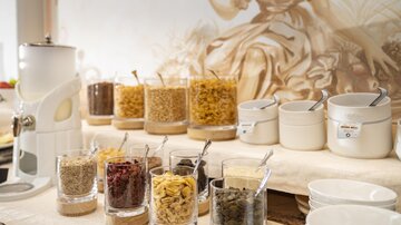 A breakfast buffet with various cereals and dried fruits in jars. In the background, a cozy mural can be seen.