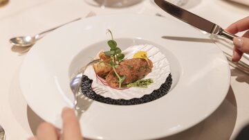 An elegant dish on a white plate, garnished with fresh herbs. The food sits on a bed of black caviar and is held by a person with cutlery.