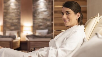 A young woman in a bathrobe sits relaxed in a modernly designed room. Soft light and wood paneling create a cozy atmosphere.