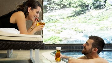 A couple relaxes by the pool and enjoys drinks. In the background, a green landscape is visible.
