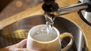 A hand holds a white cup while water flows from a faucet. In the background, a modern coffee machine is visible.