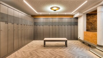 A modern changing room with gray lockers and a neutral wooden table in the center. The walls are brightly lit and the floor has an appealing pattern.