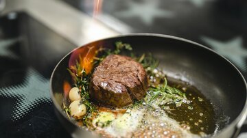 A juicy piece of meat is frying in a pan with fresh herbs and a bit of butter. A small flare adds an exciting touch to the dish.