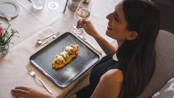 A woman is sitting at a beautifully set table, enjoying a delicious dish. She is holding a glass of wine in her hand and smiling.