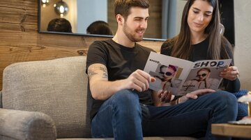 A couple is sitting on a sofa and looking at a magazine. The room has a cozy atmosphere with wooden paneling and a mirror in the background.