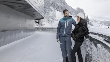 A couple stands on an ice rink in a snow-covered mountain landscape. Both are wearing winter clothing and seem to be chatting.