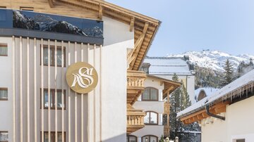 A modern building with wood cladding and a round, golden sign. In the background, snow-covered mountains can be seen.